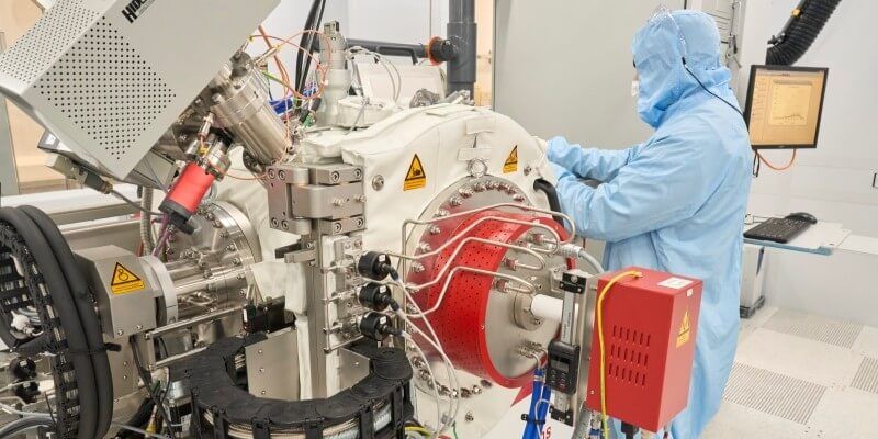 A user in a protective body suit and hood stands next to the instrument, a tall structure with a white central base and supporting wires and mechanisms.
