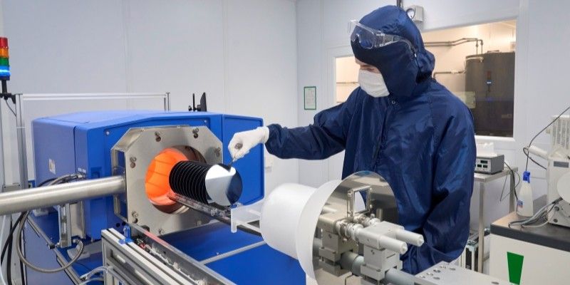 A horizontal mechanical arm loads a tray of semiconductor wafers into the furnace, a chest-height blue instrument with a round metal-edged opening and inner orange glow. A user holds one of the wafers with tweezers.