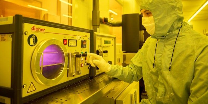 A person in light blue protective body suit and white nitrile gloves adjusts a small rectangular instrument containing purple plasma, which is visible through a circular glass door.  The room has yellow lighting.
