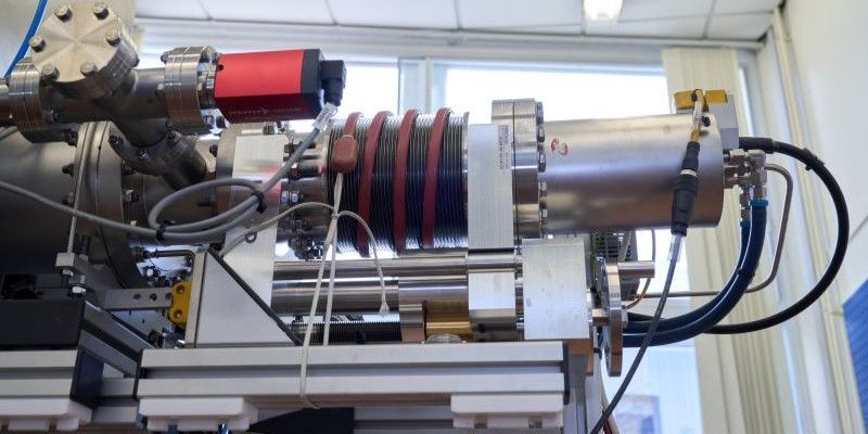 A close-up of the nanoparticle source showing a cylindrical vacuum component with integrated heating coils, electrical cabling, cooling lines and mechanical supports mounted on a laboratory frame.