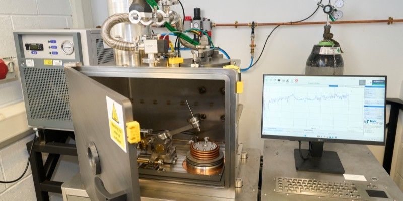 The tribometer with its chamber door open, showing a heated sample stage and contact arm inside the enclosure and connected gas and vacuum lines. There is a control computer next to the instrument displaying friction data.