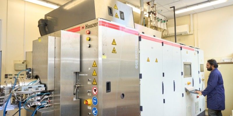 The system, a large stainless steel structure, featuring multiple interconnected metal panels, control buttons, warning labels, piping and electrical connections. A technician in a blue lab coat operates the system’s user control panel.
