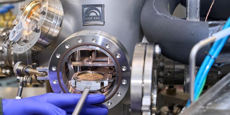 Blue gloved hands position a circular sample holder inside into the System’s an open high‑vacuum deposition chamber, with stainless steel fittings, valves and connected tubing visible around the chamber.