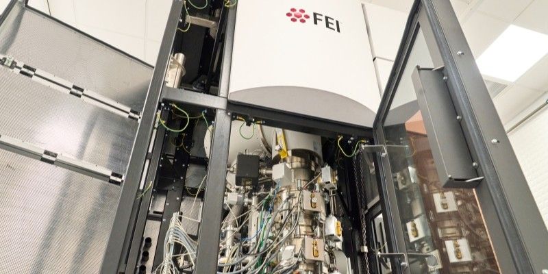 The microscope, a room-height metal and glass structure stands with its door open, revealing wiring and mechanics. The structure is branded with the FEI logo.