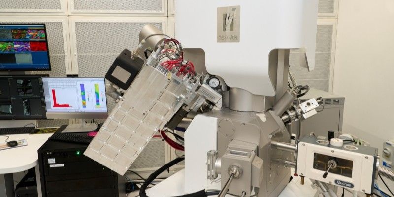 The microscope, a white plastic and metal instrument featuring the Tescan logo, next to two computer screens displaying analytical data.