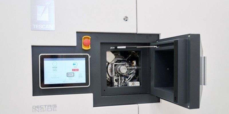A small black square compartment within the Tensor microscope, with its door open showing the loaded sample holder, a metal mechanism. The compartment sits alongside a small control panel screen, an emergency stop button and the TESCAN logo. 