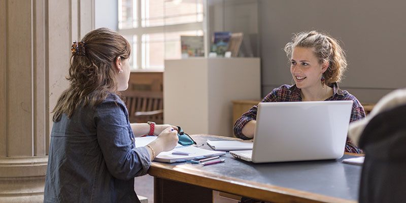 Students sat at a desk in the Parkinson Court with a laptop, chatting.