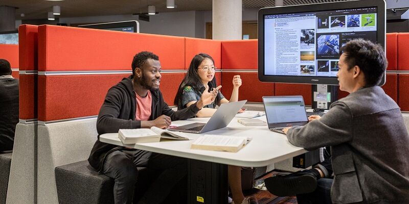 A group of students in a study booth in the library, working on laptops and a connected big screen.