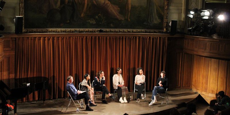 Six individuals sit on stage facing an audience in a theatre