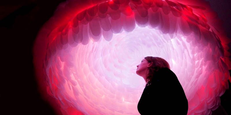 A woman looks up at a pink sculpture above her, featuring layers of pink petals.