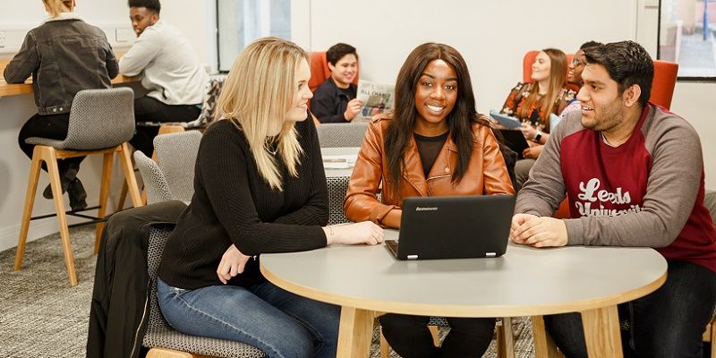 Three students sat around a laptop at a table in a busy study space with students in the background.