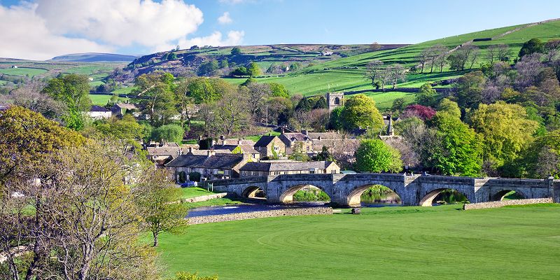 A picturesque village scene, with a bridge and a river beside a large green