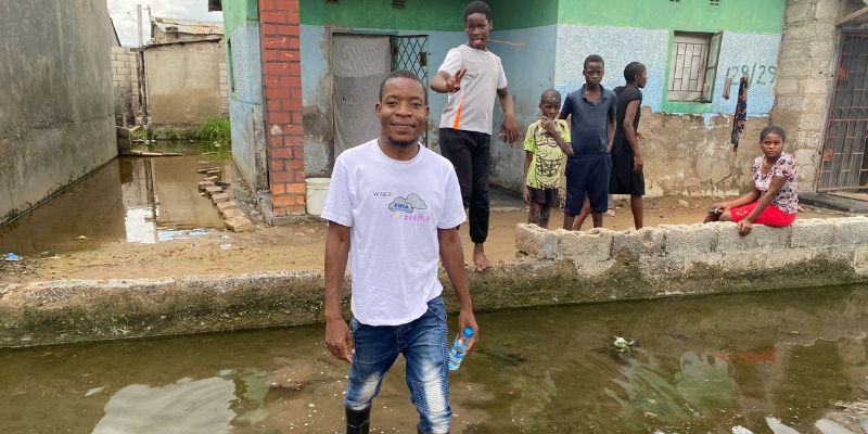 a group of people gathered around a flooded section of Kanyama village, with one individual wading through the flood waters