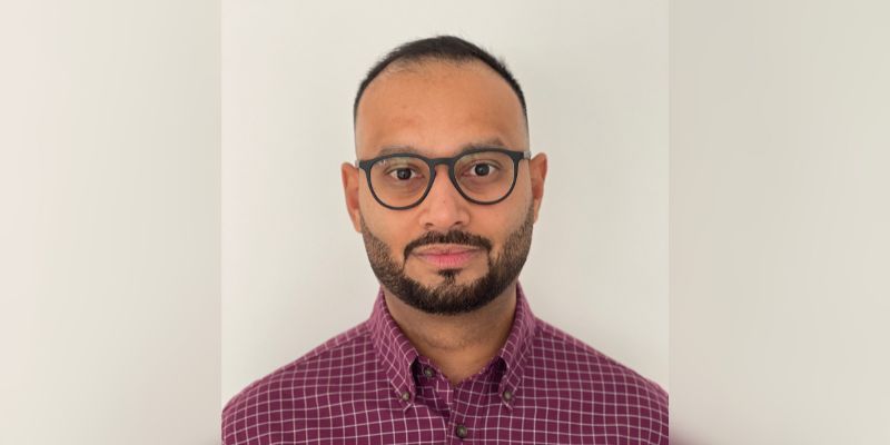 Dr Ali Malik wearing glasses and a maroon checkered button-up shirt standing against a plain light-colored background.