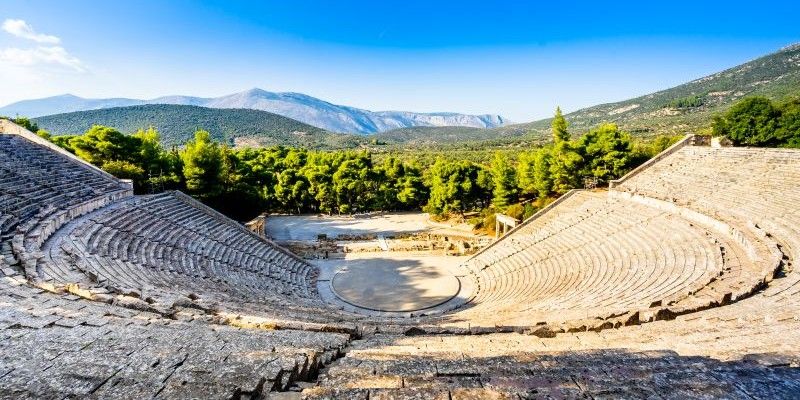 Wide view of the Ancient Theatre of Epidaurus in Greece, showing sweeping semi-circular stone seating descending to a circular stage, surrounded by green forested hills and mountains under a clear blue sky