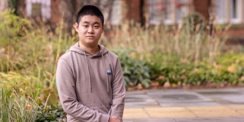 Andy sits smiling at the camera, with plants behind him and a paved pathway.