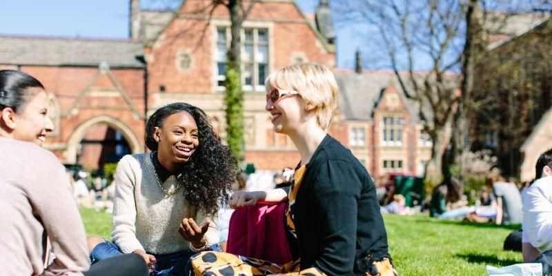 Three smiling students sitting on a patch of grass on campus in front of a redbrick building the sunshine.