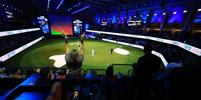An indoor arena with a seating around the outside, a green pitch in the middle and a large screen at the end. Players are standing on the green pitch and an individual stands to clap in the stands.