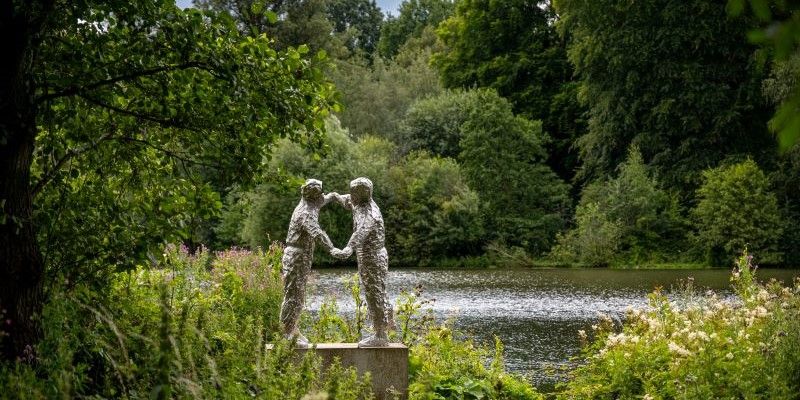 Sculpture of two people linked by their arms, positioned in a forest with a lake in the background.