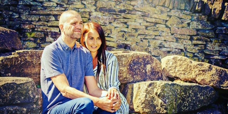 Dominic and Jessica sit in front of stone wall holding hands