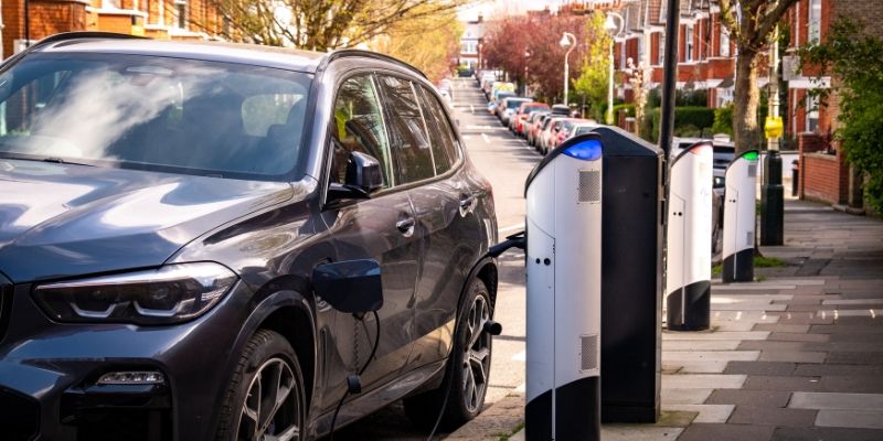 Electric car charges at a pavement charging point on a sunny suburban street. The car is in the foreground of the image, with a row of red brick houses and parked cars behind it.