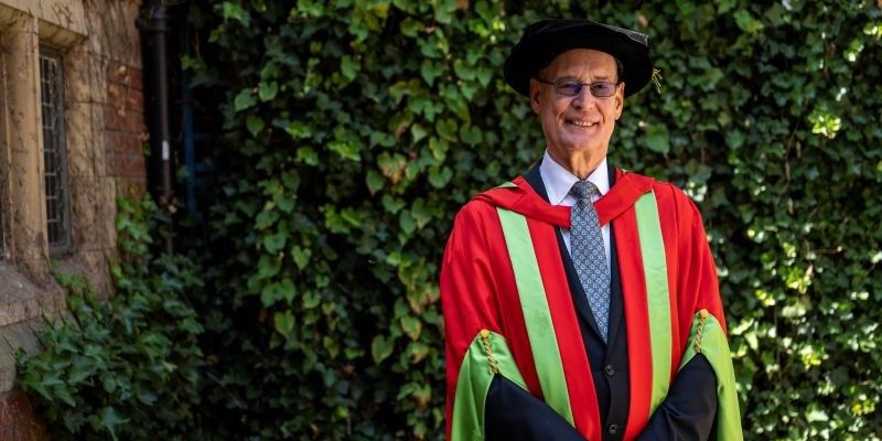 Ed Anderson stands in front of a green leafy wall and stone building, wearing the honorary graduate green and red robes. He is smiling. 