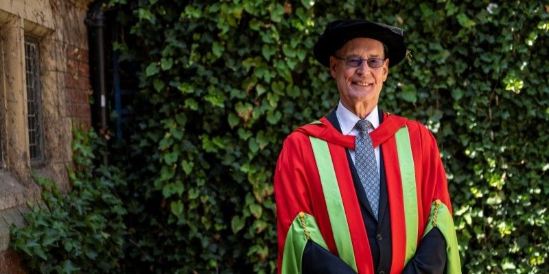 Ed Anderson CBE stands smiling at the camera in red and green graduation gowns and a black cap.