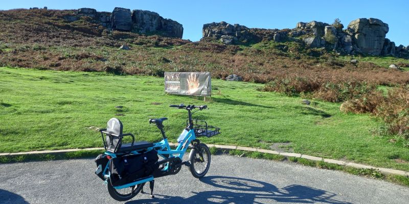 Blue electric cargo bike with a child seat and front basket parked on a paved area, set against a grassy hillside with scattered rocks and large rocky outcrops under a clear blue sky.