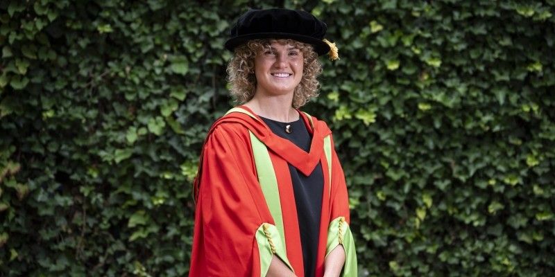 Ellie Kildunne stands smiling at the camera in red and green graduation gowns and a black cap.