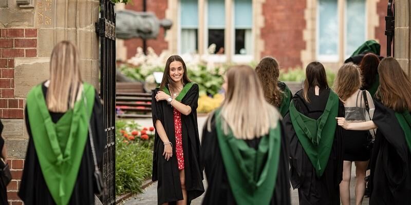 Graduates in their gowns on graduation day. One graduate is facing the camera smiling and wearing a red dress and a gown