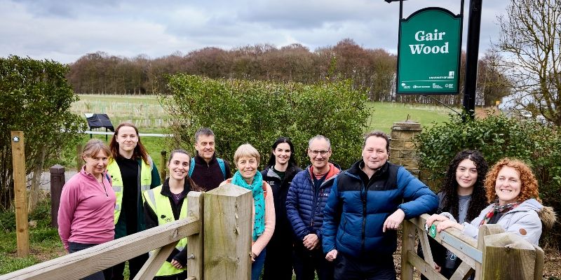 Nick Plant is leaning on the gate marking the entrance to the new pathway across Gair Wood site. He is surrounded by volunteers called Friends of Gair Wood.