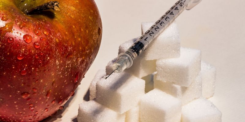 Insulin injection needle perched on cubes of sugar next to an apple.