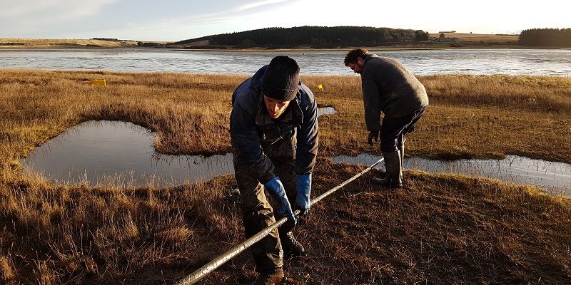Two people in winter coats use a long pole to collect core samples on wet plain with lake in background