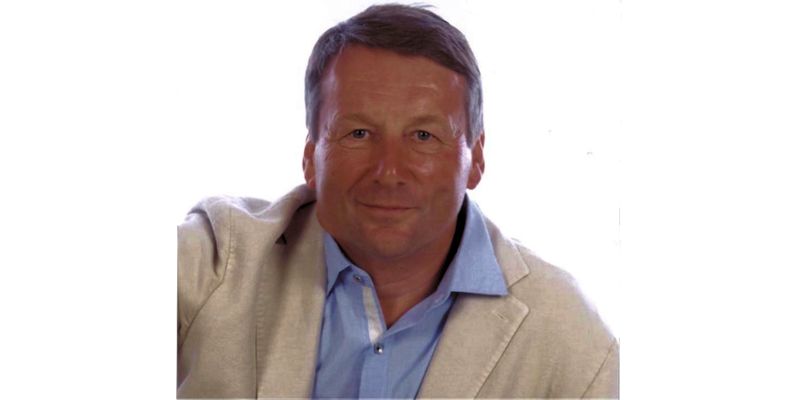 A middle-aged white man, John Barnes, smiles at camera whilst wearing a blue shirt and cream jacket.