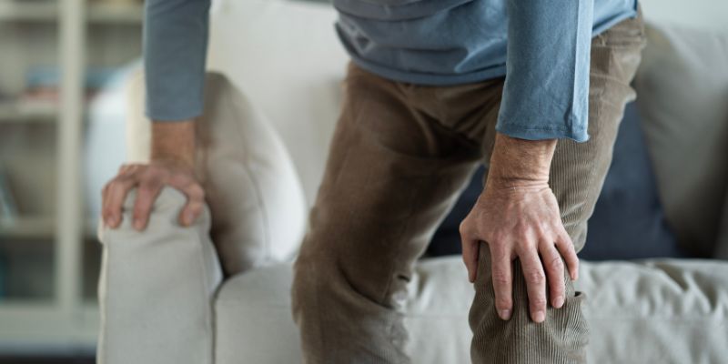Close-up of a person rising from a light-coloured sofa, leaning forward and gripping their knee with one hand while using the other hand to push off the armrest, suggesting knee pain or stiffness. This is in a home living room setting.