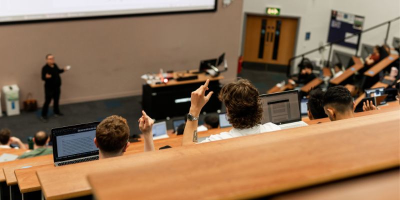 Students using laptops and raising their hands in a tiered lecture theatre.