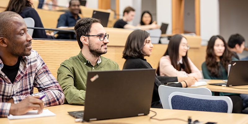 Several students in a lecture theatre listening to a presentation. There are laptops and notebooks on the study surfaces.