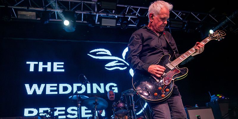 David Gedge plays guitar on stage. Behind him, a drummer is drumming, and in the backdrop a fluorescent sign reads "The Wedding Present"