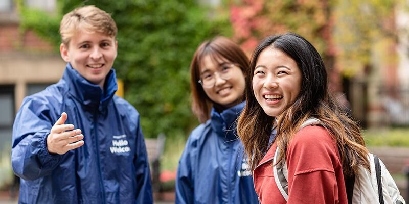 Two student ambassadors in blue coats and a student with a red jacket and grey backpack are facing the camera and smiling