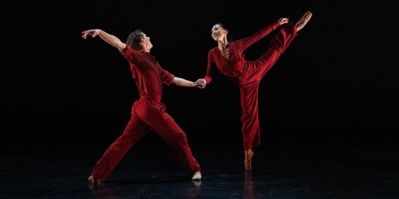 Two ballet dancers dressed in red pose against a black background, one in an arabesque