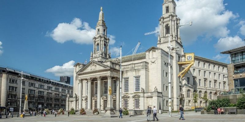 A grand stone civic building with tall twin clock towers, large arched windows, and a columned entrance stands under a bright blue sky with scattered clouds. The open square in front of the building has a few people walking across wide paved area.