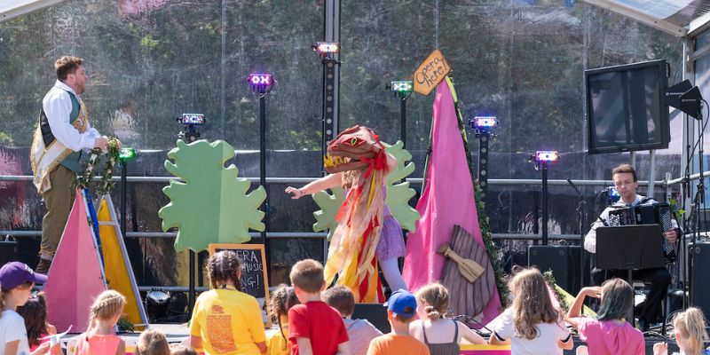 Children gather around a stage to watch a woman with a dragon puppet on the right and a man dressed as a prince on the left. A sign in the back says 'Opera here!'.