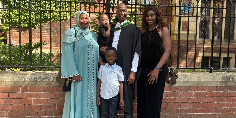 Musa Cham smiles outside the Great Hall at the University of Leeds, wearing his graduation robes and carrying his baby daughter as his sister, wife and son stand around him.
