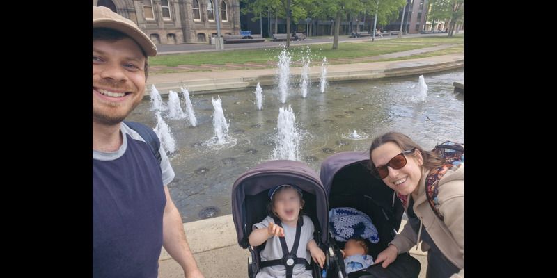 A family of two adult, a toddler and a baby smile at the camera in a selfie taken in front of a fountain in a public park