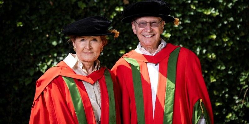 Peter and Susan Cheney stand smiling in red and green graduation robes and black caps.