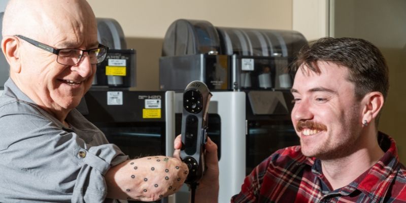 Two people in a lab. One uses a handheld scanner to scan the other person's upper arm residual limb, which has small sensors stuck on it. There are 3D printers in the background.