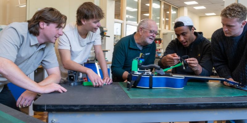 Five people gather around a workbench in a workshop, examining a device strapped to a blue guitar, with various tools and components. There is lab equipment and windows in the background.