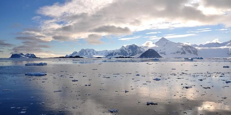 Image across a calm sea with floating ice debris to a coastline of ice and snow covered mountains. This is a the Antarctic Peninsula.