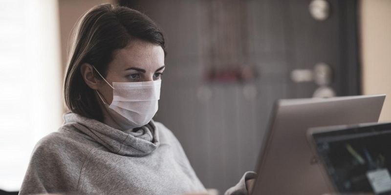 The image shows a woman wearing a mask sitting at a computer