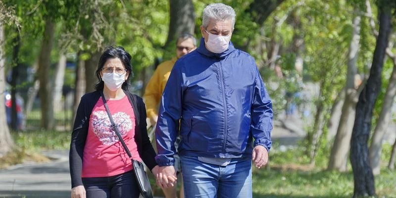 The images shows a couple, holding hands, walking through a park. They are both wearing face masks.